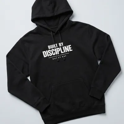 Bold typography shirt with the phrase 'BUILT BY DISCIPLINE DAY BY DAY' in black on white — printed on a unisex hoodie