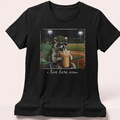 Vintage distressed print of a playful raccoon with the text 'i live here now' — printed on a women's shirt